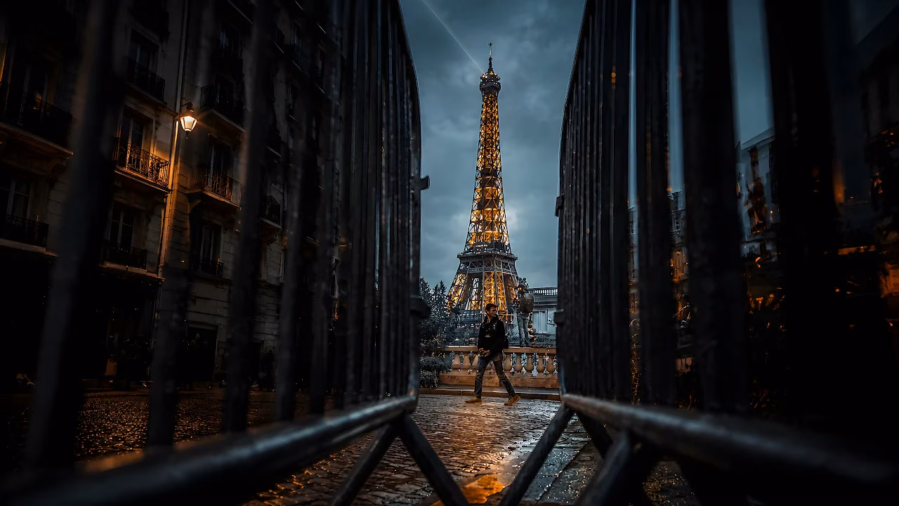 Illuminated Eiffel Tower framed between dark metal security fences with a person walking on cobblestone below.