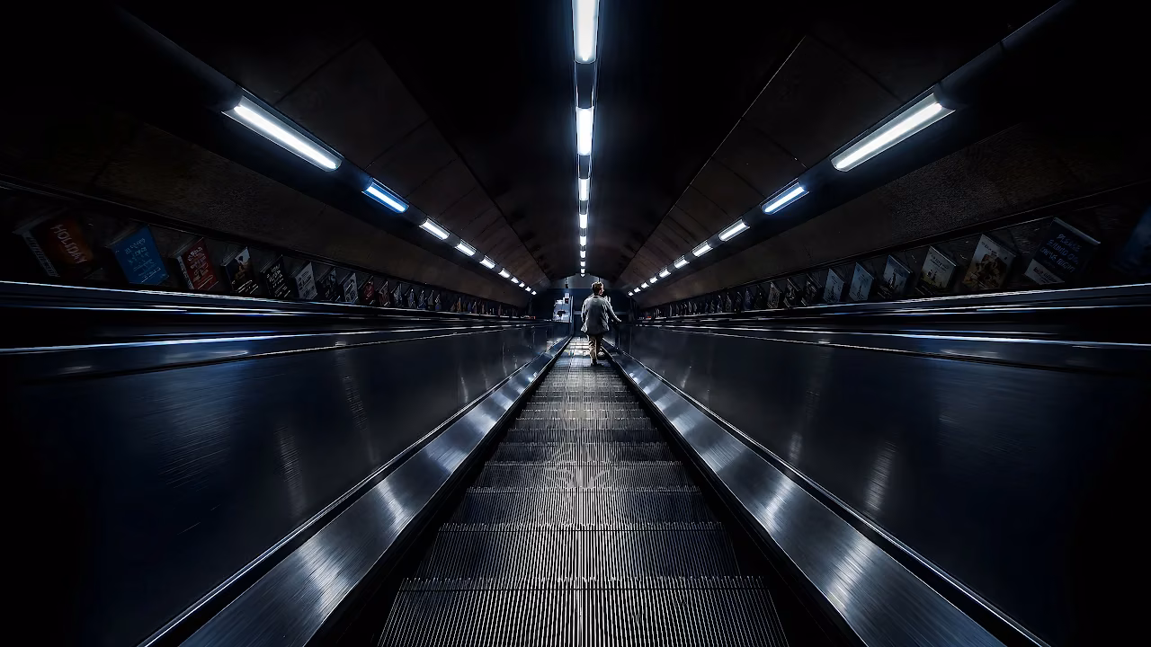 Long metal escalator ascending through a dark curved subway tunnel with fluorescent ceiling lights.