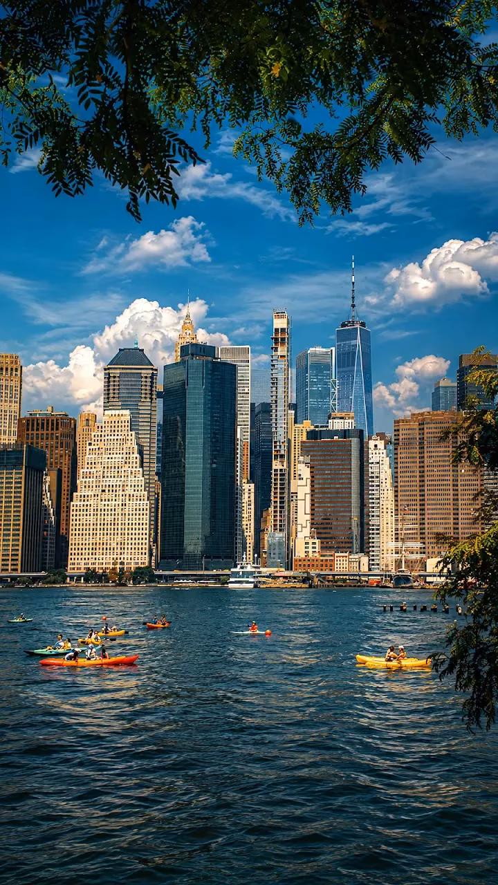 Download NYC Skyline and Kayakers in Summer 4K Mobile Wallpaper Wallpaper in Mobile HD Kayakers paddle across blue water in front of the towering Lower Manhattan skyline under a partly cloudy sky, framed by tree leaves.