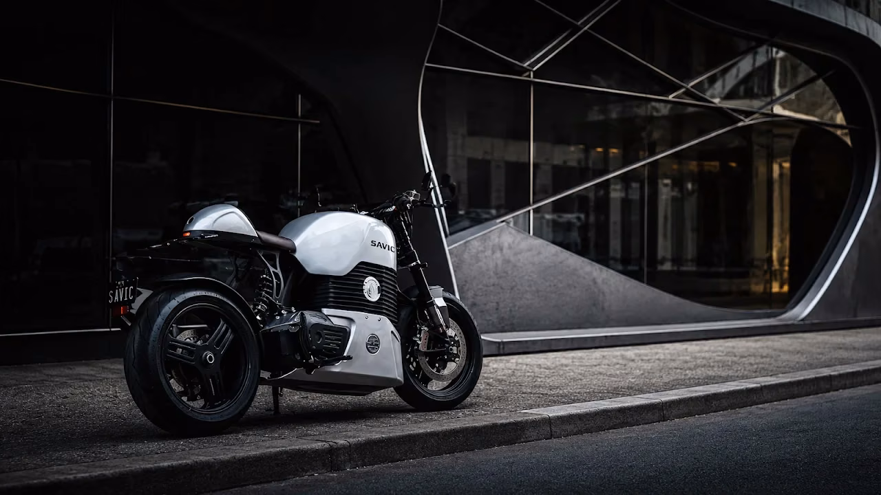Download Savic Electric Motorcycle Modern Architecture 4K Wallpaper in 4K UHD White Savic electric motorcycle parked on a concrete curb in front of an architectural modern building with large curved glass.