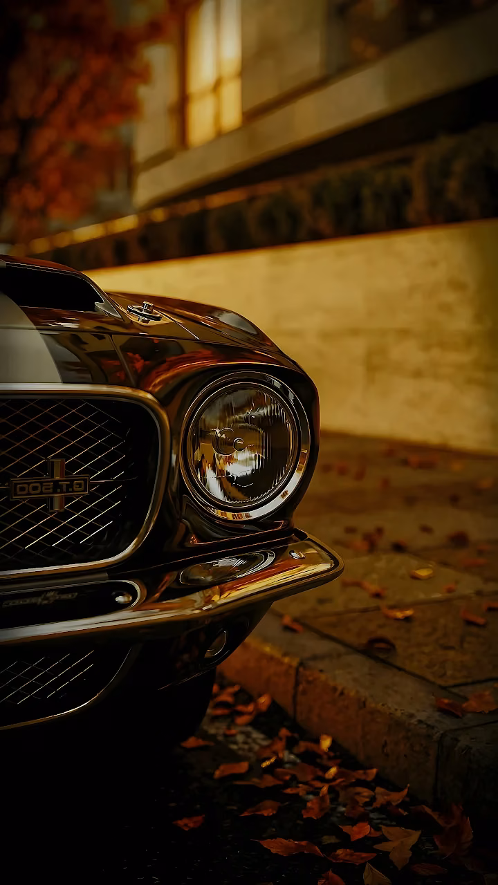 Round headlight and front chrome grille of a classic muscle car with fallen autumn leaves on pavement.