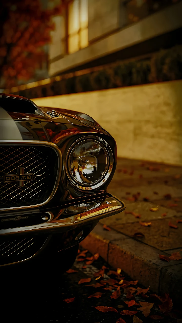 Download Shelby GT500 Chrome Headlight Autumn Leaves 4K Wallpaper in Mobile HD Round headlight and front chrome grille of a classic muscle car with fallen autumn leaves on pavement.