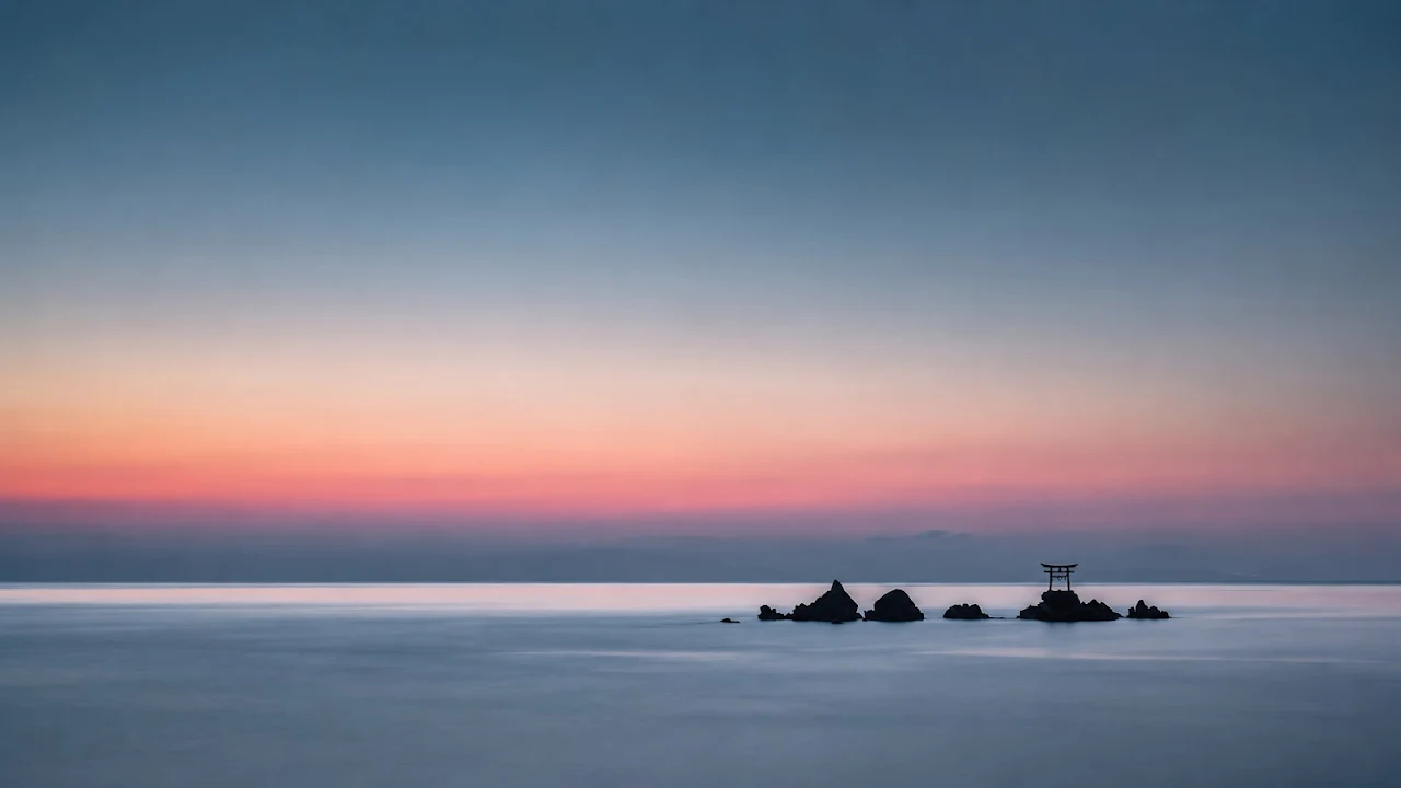 Small black torii gate perched on offshore rocks in calm blue ocean water beneath a pink sunset gradient sky.