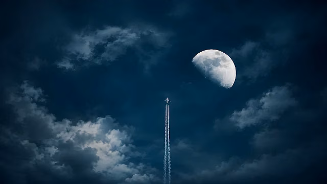 A passenger jet flies upward leaving a white contrail beside the moon in a dark night sky. Airplane ascending vertically with a white contrail next to a bright moon in a dark cloudy night sky.