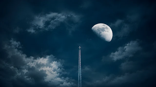 A passenger jet flies upward leaving a white contrail beside the moon in a dark night sky. Airplane ascending vertically with a white contrail next to a bright moon in a dark cloudy night sky.