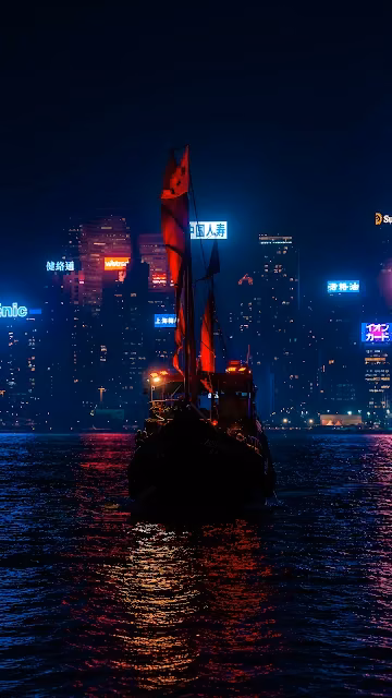 Traditional Chinese junk boat with illuminated red sails on dark harbour water facing glowing city skyscrapers.
