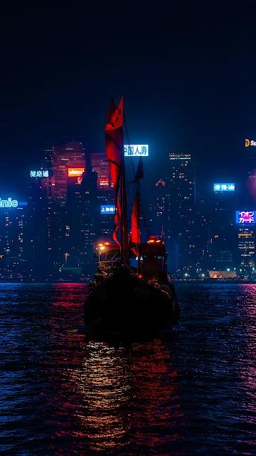Traditional Chinese junk boat with illuminated red sails on dark harbour water facing glowing city skyscrapers.