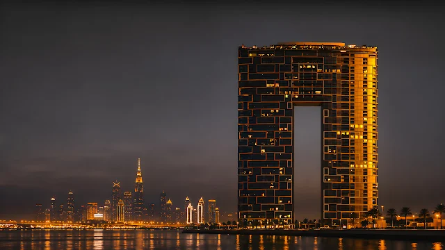 Illuminated Address Beach Resort tower stands over water with the Dubai city skyline glowing at night. Address Beach Resort arch skyscraper illuminated at night across the water with the Dubai skyline.