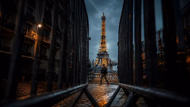 Illuminated Eiffel Tower stands behind metal street barriers during evening in Paris. Illuminated Eiffel Tower framed between dark metal security fences with a person walking on cobblestone below.