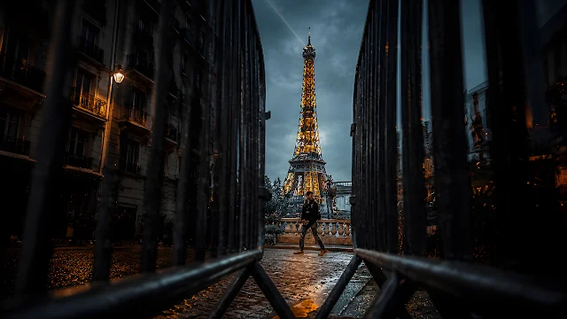 Illuminated Eiffel Tower stands behind metal street barriers during evening in Paris. Illuminated Eiffel Tower framed between dark metal security fences with a person walking on cobblestone below.