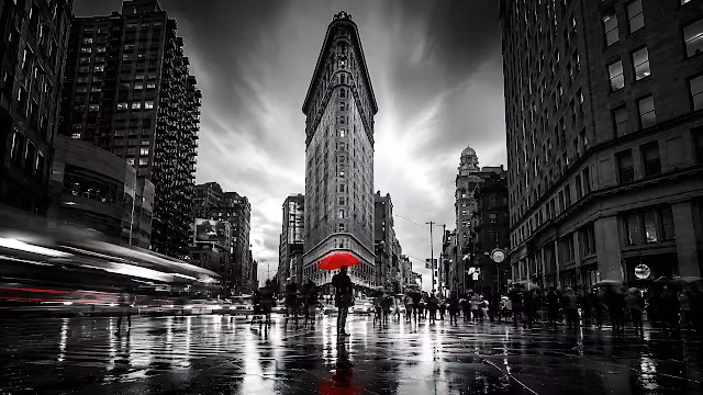 Monochrome view of the Flatiron Building in New York City with a person holding a red umbrella on wet pavement.