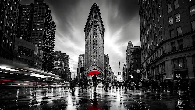 Pedestrian with a red umbrella stands before the Flatiron Building on wet New York streets. Monochrome view of the Flatiron Building in New York City with a person holding a red umbrella on wet pavement.