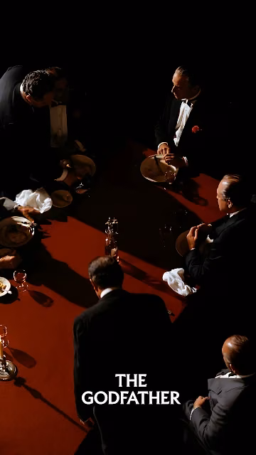 Overhead view of mafia figures in tuxedos seated around a red table under dramatic lighting with The Godfather logo. High-angle view of men in tuxedos seated around a red dining table in dark shadow with The Godfather text.