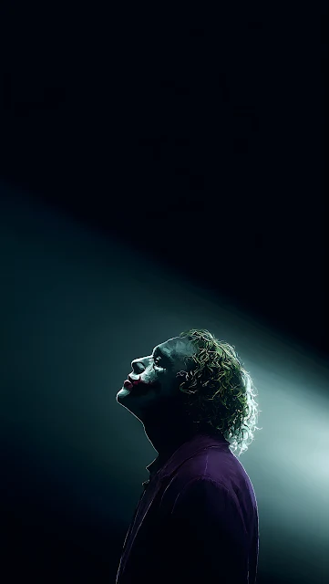 The Joker looks upward into a stark cinematic beam of light against pure shadow. Side profile of The Joker looking up toward a bright spotlight against a dark background.
