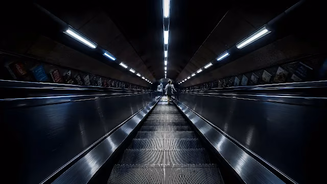 Long underground subway escalator ascends under linear ceiling lights in a metro station. Long metal escalator ascending through a dark curved subway tunnel with fluorescent ceiling lights.