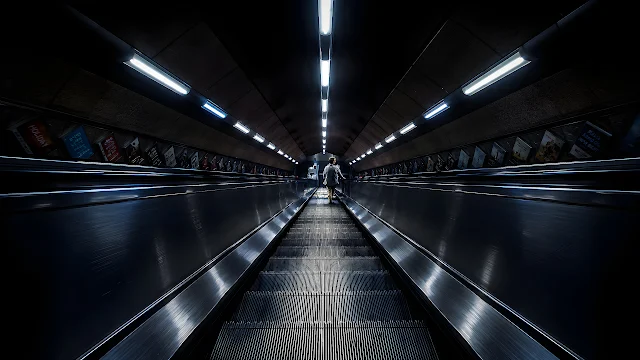 Long underground subway escalator ascends under linear ceiling lights in a metro station. Long metal escalator ascending through a dark curved subway tunnel with fluorescent ceiling lights.