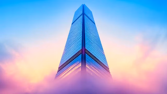Modern glass skyscraper rises through pastel pink sunset clouds into clear blue sky. Low-angle view of a tall modern glass skyscraper rising through pink and purple clouds at sunset.