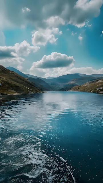 Calm blue mountain lake with boat wake under a bright sky with isolated cumulus clouds. Deep blue mountain lake with boat wake in the foreground, flanked by grassy hills under a cloudy blue sky.