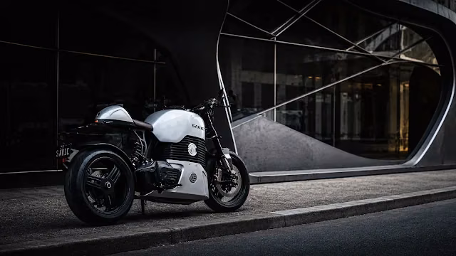 White Savic electric motorcycle parked on a concrete curb in front of an architectural modern building with large curved glass.