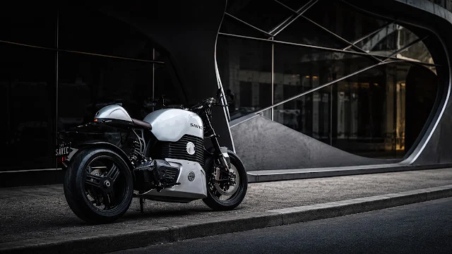 A custom white Savic electric motorcycle parked in front of a futuristic glass building. White Savic electric motorcycle parked on a concrete curb in front of an architectural modern building with large curved glass.