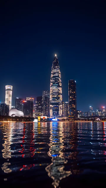 The illuminated China Resources Tower and Shenzhen skyline reflect over calm bay waters at night. Shenzhen skyline and illuminated bamboo shoot tower reflecting across water at night.