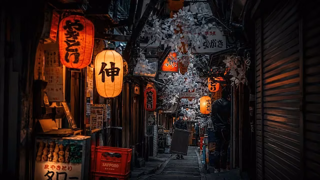 Narrow Tokyo alleyway at night illuminated by glowing red and orange paper lanterns with a pedestrian walking past izakaya storefronts.