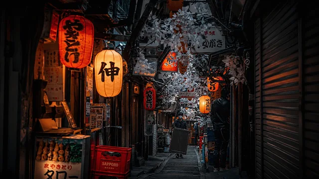 Glowing orange paper lanterns light a narrow Shinjuku alleyway framed by artificial cherry blossoms. Narrow Tokyo alleyway at night illuminated by glowing red and orange paper lanterns with a pedestrian walking past izakaya storefronts.