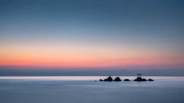 Small black torii gate perched on offshore rocks in calm blue ocean water beneath a pink sunset gradient sky.