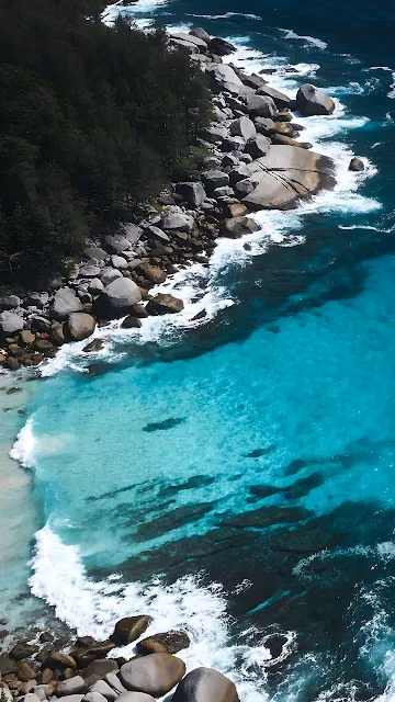 Aerial view of turquoise ocean waves breaking against coastal granite boulders. Top-down view of clear turquoise water and surf breaking on smooth rocky coastline.