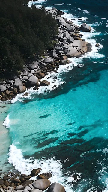 Top-down view of clear turquoise water and surf breaking on smooth rocky coastline.